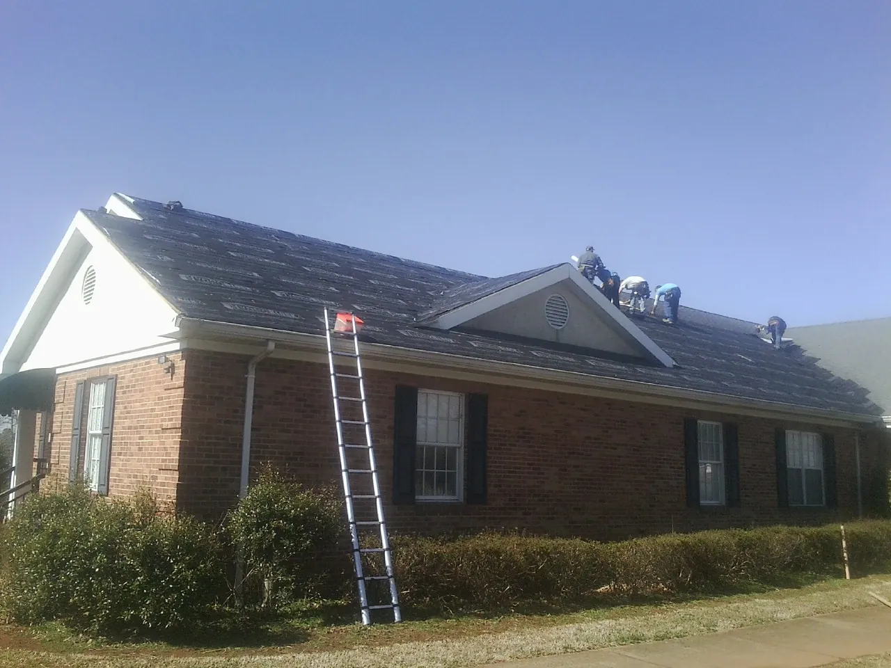 Crew tearing shingles off a brick ranch roof with a ladder set — Metal Roof Repair work by our crew in Amite