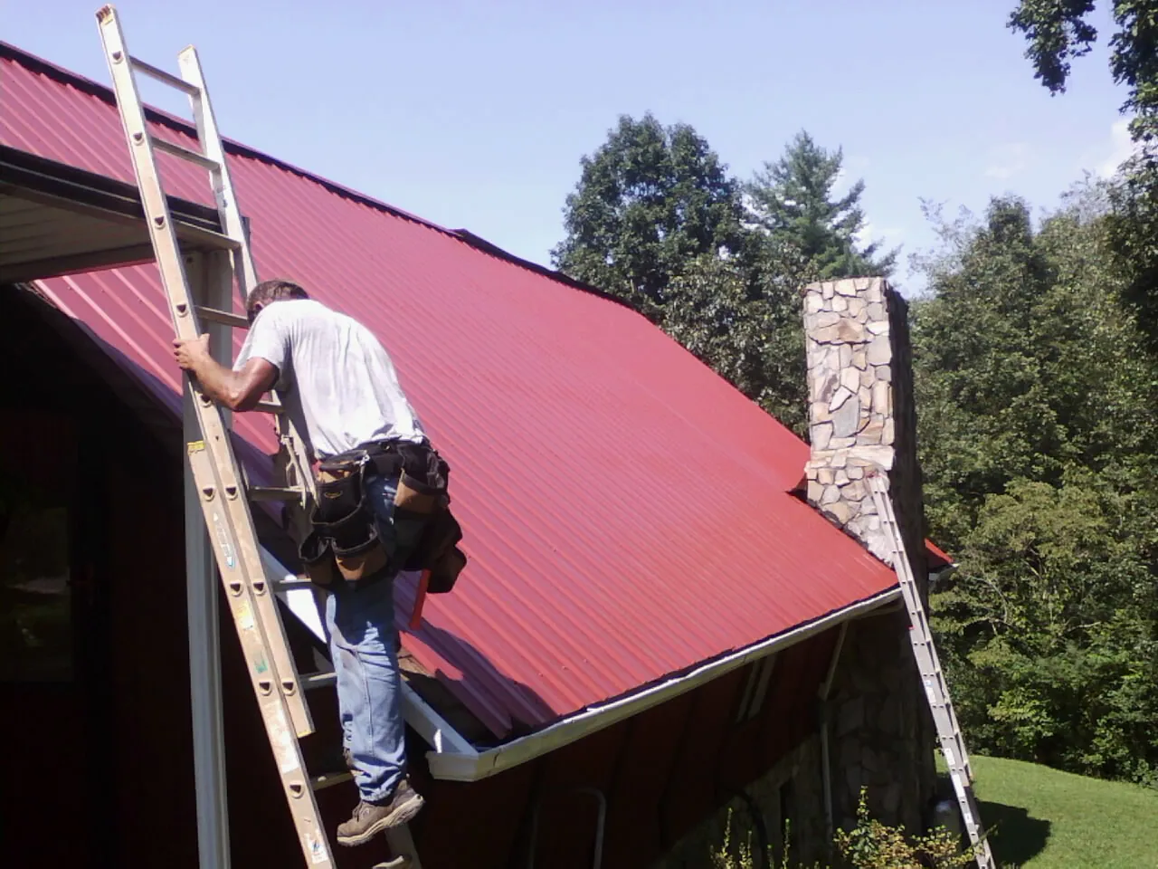 Roofer on a ladder beside a red metal roof and stone chimney — Soffit & Fascia Repair by Roofing Brothers in Amite